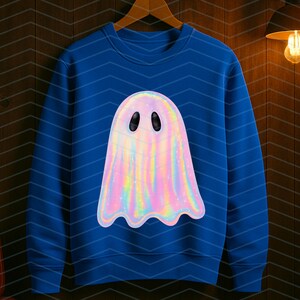 Puede incluir: Una sudadera azul con un dise&ntilde;o de fantasma hologr&aacute;fico. El fantasma tiene un acabado nacarado e iridiscente con ojos ovalados negros. La sudadera est&aacute; colgada de una percha de madera contra una pared de paneles de madera oscura.