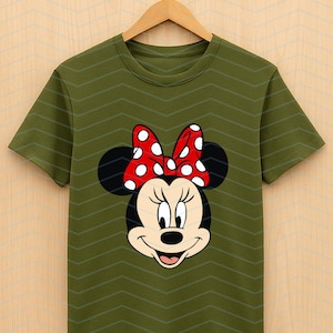 May include: Olive green t-shirt featuring a Minnie Mouse graphic. The graphic shows Minnie's face with a red bow with white polka dots. The shirt is hanging on a wooden hanger.