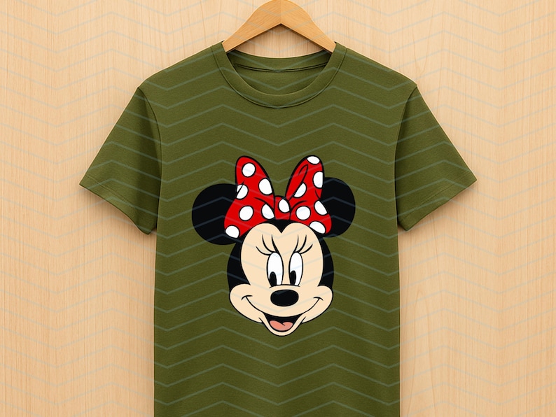 May include: Olive green t-shirt featuring a Minnie Mouse graphic. The graphic shows Minnie's face with a red bow with white polka dots. The shirt is hanging on a wooden hanger.