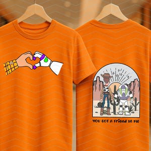 Pu&ograve; includere: T-shirt arancione con un cuore formato da una mano umana e una mano robotica. Il retro della maglietta presenta una scena desertica con due personaggi dei cartoni animati e la scritta "YOU GOT A FRIEND IN ME".