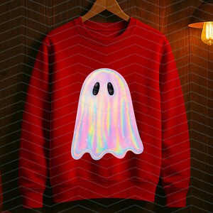 Puede incluir: Una sudadera roja con un gran gr&aacute;fico de fantasma iridiscente. El fantasma tiene una apariencia nacarada, similar al arco&iacute;ris, con ojos ovalados negros. La sudadera est&aacute; colgada en una percha de madera contra una pared de paneles de madera oscura.