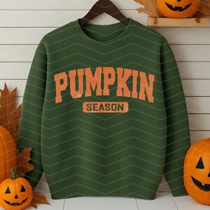 May include: Green sweatshirt with the words "PUMPKIN SEASON" in orange block letters. The sweatshirt is hanging on a wooden hanger. Pumpkins and autumn leaves are arranged around the sweatshirt, creating an autumnal theme.