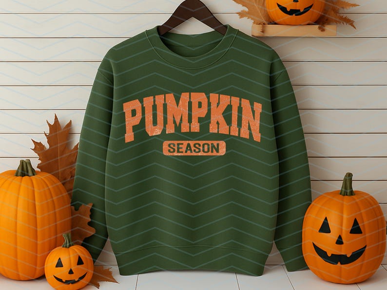 May include: Green sweatshirt with the words "PUMPKIN SEASON" in orange block letters. The sweatshirt is hanging on a wooden hanger. Pumpkins and autumn leaves are arranged around the sweatshirt, creating an autumnal theme.