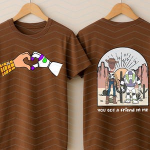 Pu&ograve; includere: T-shirt marrone con un cuore fatto di mani dei cartoni animati, una con un motivo giallo e marrone e l'altra bianca con accenti viola e verdi. Il retro della maglietta mostra Woody e Buzz Lightyear con la scritta "you got a friend in me".