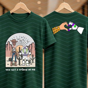 Pu&ograve; includere: T-shirt verde scuro con una grafica di due personaggi dei cartoni animati in un paesaggio desertico e la scritta "you got a friend in me". Il retro della maglietta mostra delle mani che formano un cuore. La maglietta &egrave; appesa a una gruccia di legno.