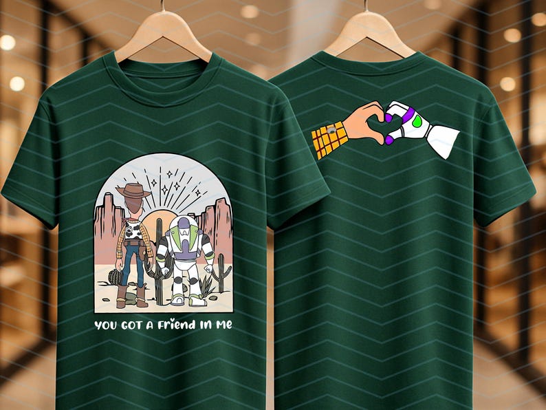 Pu&ograve; includere: T-shirt verde scuro con una grafica di due personaggi dei cartoni animati in un paesaggio desertico e la scritta "you got a friend in me". Il retro della maglietta mostra delle mani che formano un cuore. La maglietta &egrave; appesa a una gruccia di legno.