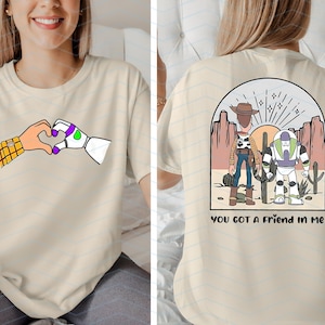 Pu&ograve; includere: T-shirt beige con un cuore formato da mani di cartoni animati, una con una manica a quadri e l'altra robotica. Il retro della maglietta mostra una scena desertica con due personaggi e la scritta "YOU GOT A FRIEND IN ME".