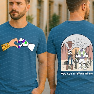 Pu&ograve; includere: T-shirt blu teal con una grafica di due mani che formano un cuore, una umana e una robotica. Il retro della maglietta mostra Woody e Buzz Lightyear in una scena desertica con la scritta "you got a friend in me."