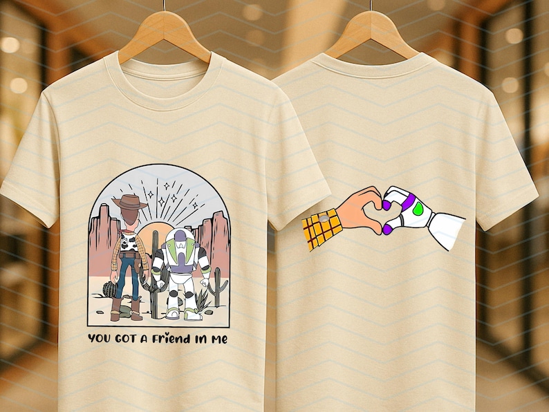 Pu&ograve; includere: T-shirt beige con un'illustrazione a cartoni animati di due personaggi, un cowboy e un ranger spaziale. La parte anteriore della maglietta ha la scritta "YOU GOT A FRIEND IN ME". Il retro della maglietta mostra mani che formano un cuore.