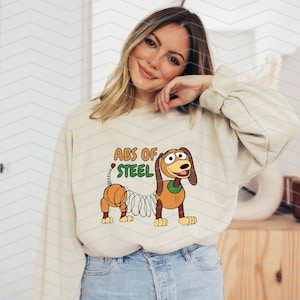 May include: Cream-colored sweatshirt featuring a cartoon illustration of a dachshund with a spring body. The text "ABS OF STEEL" is above the dog. The dog is brown, orange, and green. The model is wearing blue jeans.