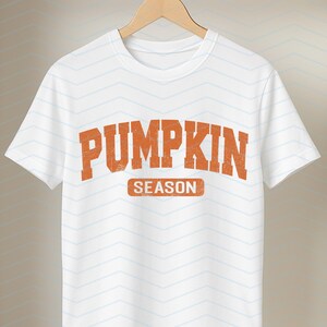 May include: White t-shirt with the words "PUMPKIN SEASON" in orange, distressed lettering. The text is centered on the shirt. The shirt is hanging on a wooden hanger against a white background with a subtle chevron pattern.