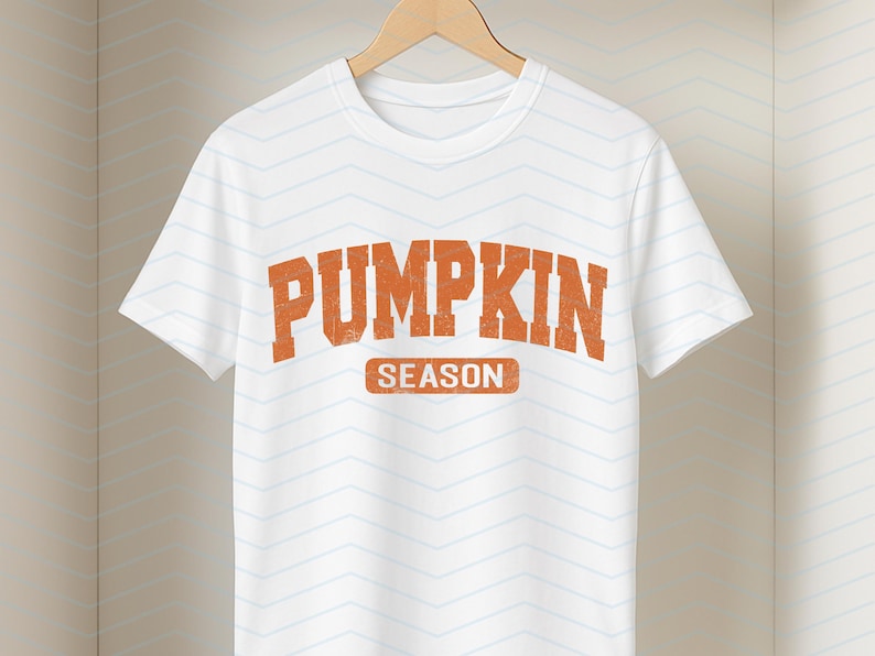 May include: White t-shirt with the words "PUMPKIN SEASON" in orange, distressed lettering. The text is centered on the shirt. The shirt is hanging on a wooden hanger against a white background with a subtle chevron pattern.