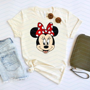 May include: Cream-colored t-shirt featuring a Minnie Mouse graphic with a red polka-dot bow. The shirt is paired with denim shorts, sunglasses, and a green wallet. The design is perfect for Disney fans.