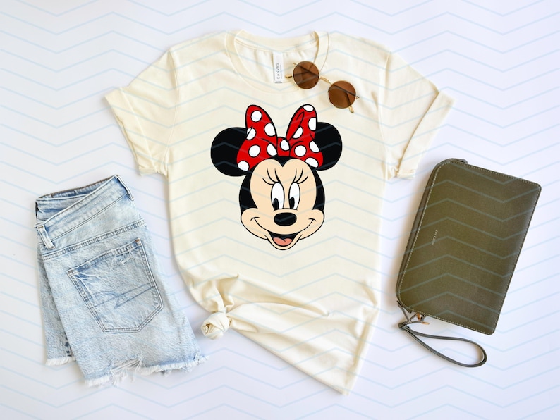 May include: Cream-colored t-shirt featuring a Minnie Mouse graphic with a red polka-dot bow. The shirt is paired with denim shorts, sunglasses, and a green wallet. The design is perfect for Disney fans.