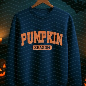 May include: A navy blue sweatshirt with the words "PUMPKIN SEASON" in orange block letters. The sweatshirt is hanging on a wooden hanger. The background has a chevron pattern and lit jack-o'-lanterns and candles.
