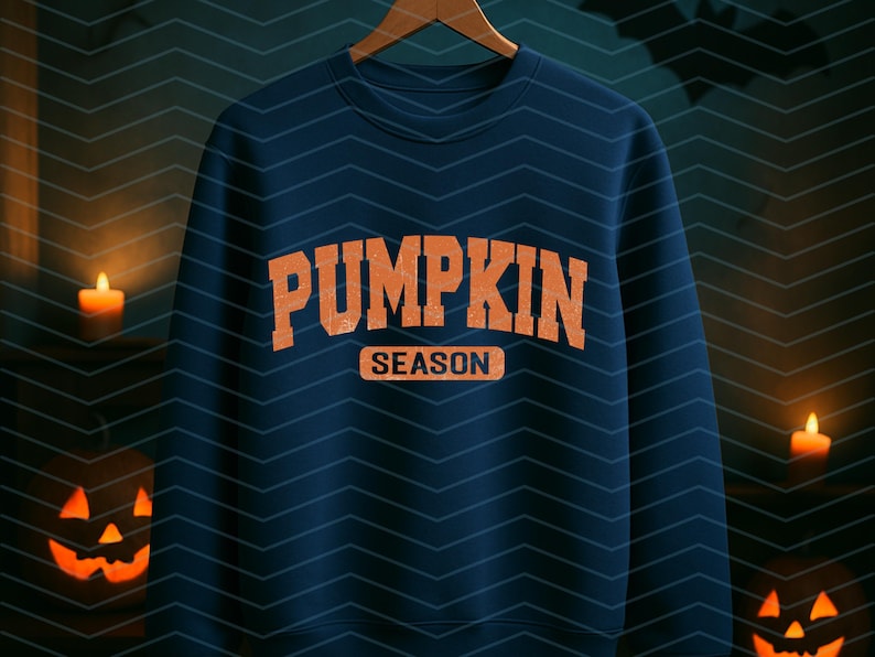 May include: A navy blue sweatshirt with the words "PUMPKIN SEASON" in orange block letters. The sweatshirt is hanging on a wooden hanger. The background has a chevron pattern and lit jack-o'-lanterns and candles.