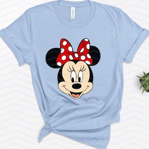 May include: Light blue t-shirt featuring a graphic of Minnie Mouse's face. Minnie's face is black and white with a red bow with white polka dots. The shirt is tied at the bottom.