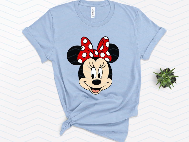 May include: Light blue t-shirt featuring a graphic of Minnie Mouse's face. Minnie's face is black and white with a red bow with white polka dots. The shirt is tied at the bottom.