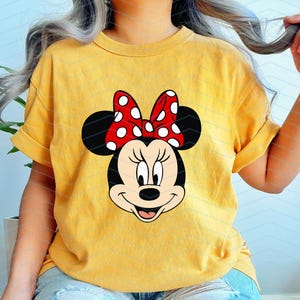 May include: Yellow t-shirt featuring a Minnie Mouse graphic. The graphic shows Minnie's face with a red bow with white polka dots. The shirt has rolled-up sleeves and is worn by a person.