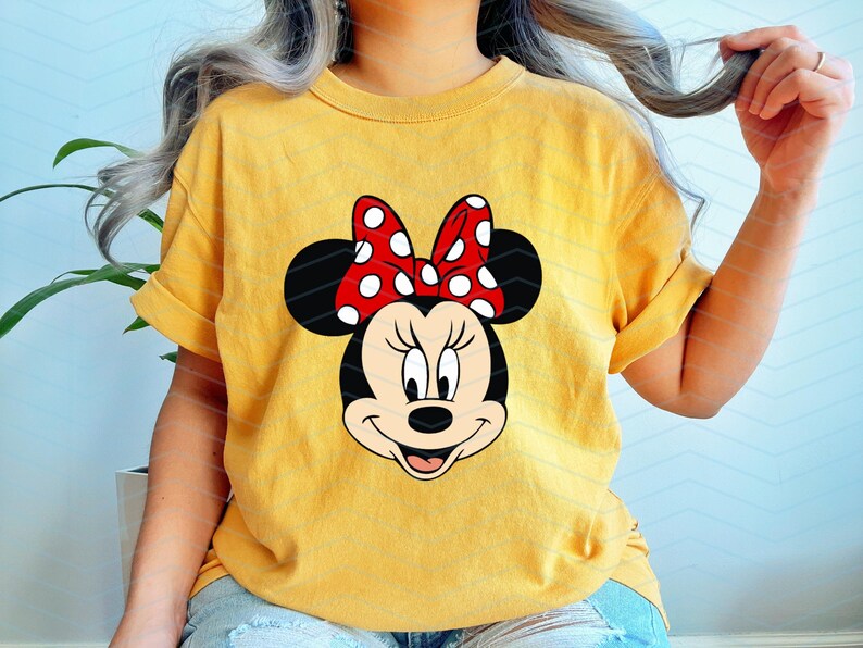 May include: Yellow t-shirt featuring a Minnie Mouse graphic. The graphic shows Minnie's face with a red bow with white polka dots. The shirt has rolled-up sleeves and is worn by a person.
