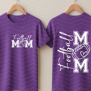 May include: Purple t-shirt with a white graphic that says "Football Mom" with a football and heart design. The back of the shirt has the words "A Football Mom" with a football and heart design.