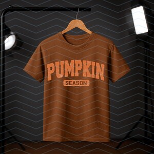 May include: A brown t-shirt with the words "PUMPKIN SEASON" in orange block letters. The shirt is hanging on a wooden hanger against a dark background with studio lights. The design has a distressed, vintage appearance.