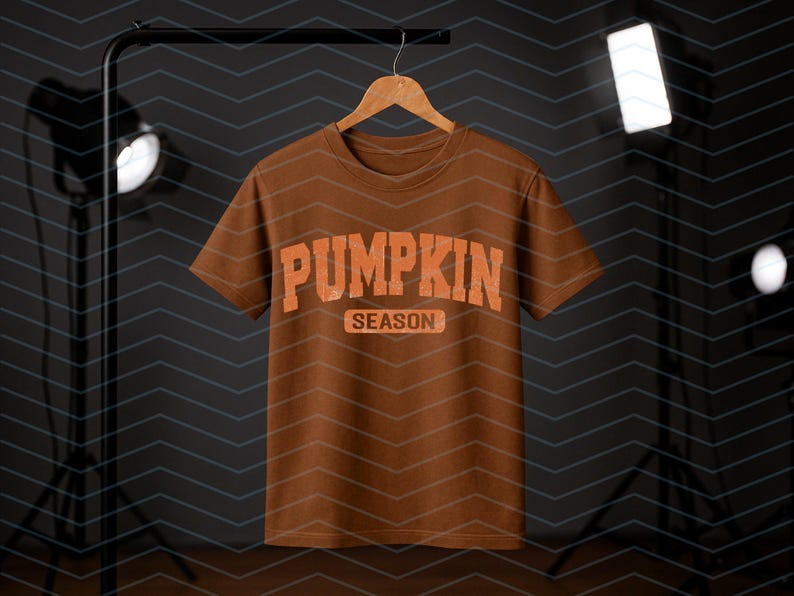 May include: A brown t-shirt with the words "PUMPKIN SEASON" in orange block letters. The shirt is hanging on a wooden hanger against a dark background with studio lights. The design has a distressed, vintage appearance.