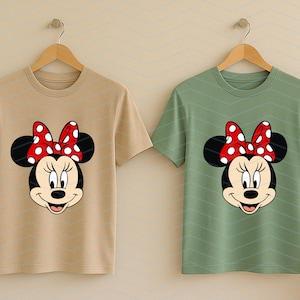 May include: Two t-shirts, one tan and one green, each featuring a Minnie Mouse graphic. The graphic shows Minnie's face with a red polka-dot bow. The shirts are short-sleeved and hanging on wooden hangers.