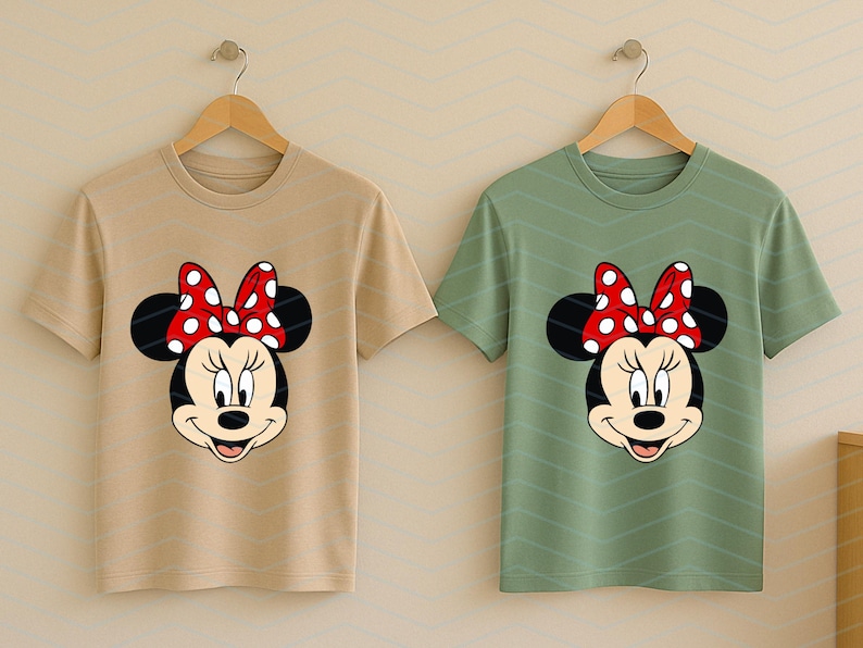 May include: Two t-shirts, one tan and one green, each featuring a Minnie Mouse graphic. The graphic shows Minnie's face with a red polka-dot bow. The shirts are short-sleeved and hanging on wooden hangers.