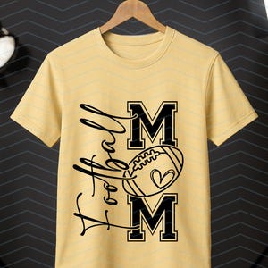 May include: A light yellow t-shirt with the words "Football Mom" in black script and block letters. A football with a heart is in the center. The shirt is hanging on a wooden hanger.
