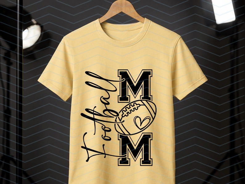 May include: A light yellow t-shirt with the words "Football Mom" in black script and block letters. A football with a heart is in the center. The shirt is hanging on a wooden hanger.