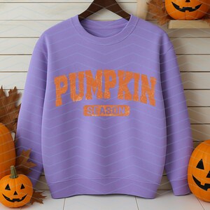 May include: A lilac sweatshirt with the words "PUMPKIN SEASON" in orange lettering. The sweatshirt is hanging on a wooden hanger. Pumpkins of various sizes are arranged around the sweatshirt, creating an autumnal theme.