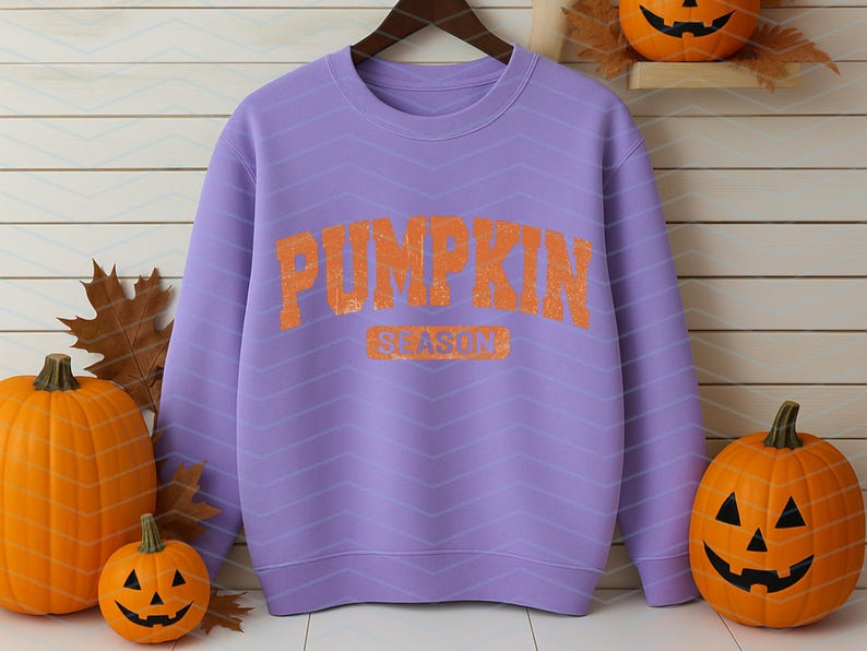 May include: A lilac sweatshirt with the words "PUMPKIN SEASON" in orange lettering. The sweatshirt is hanging on a wooden hanger. Pumpkins of various sizes are arranged around the sweatshirt, creating an autumnal theme.