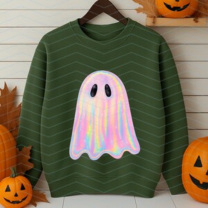 Puede incluir: Sudadera verde con un dise&ntilde;o de fantasma hologr&aacute;fico. El fantasma es de color arco&iacute;ris pastel con ojos ovalados negros. La sudadera se muestra en una percha de madera, con calabazas y hojas de oto&ntilde;o en el fondo. Un tema de Halloween.
