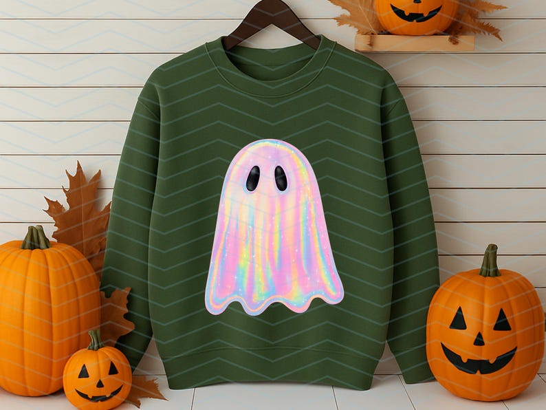 Puede incluir: Sudadera verde con un dise&ntilde;o de fantasma hologr&aacute;fico. El fantasma es de color arco&iacute;ris pastel con ojos ovalados negros. La sudadera se muestra en una percha de madera, con calabazas y hojas de oto&ntilde;o en el fondo. Un tema de Halloween.