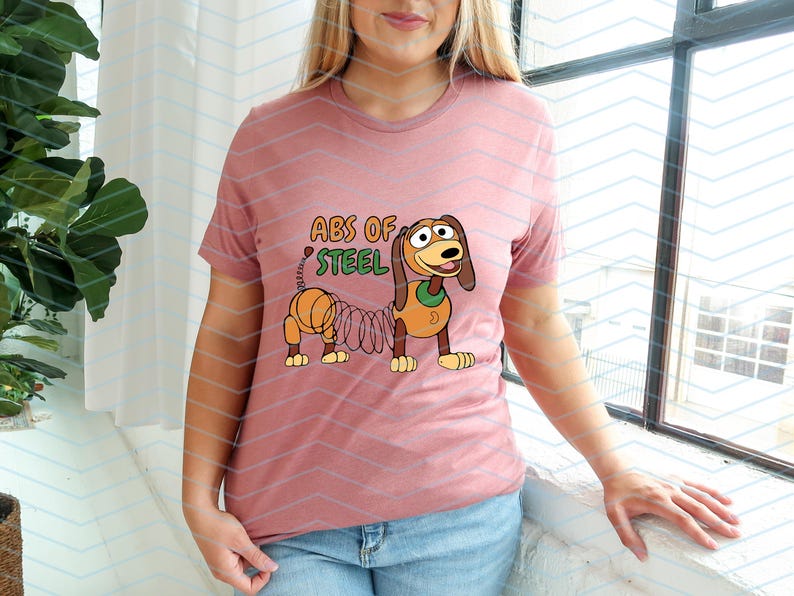 May include: A dusty rose t-shirt featuring a cartoon dog with a spring-like body and the text "ABS OF STEEL" in green and orange. The dog has brown and tan coloring with a green collar.