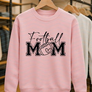 May include: A light pink sweatshirt with the words "Football Mom" in black script and block letters. The "MOM" letters frame a football with a heart. The sweatshirt is displayed on a wooden hanger with other sweatshirts in the background.