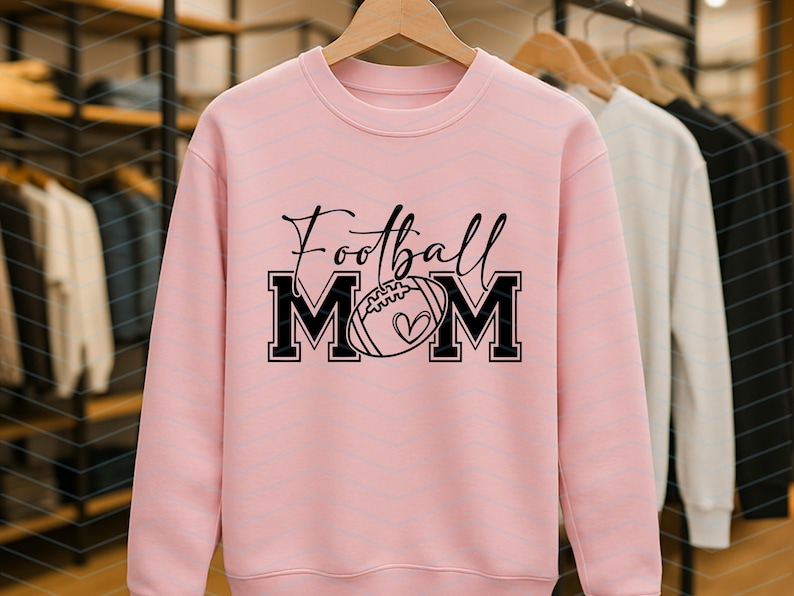 May include: A light pink sweatshirt with the words "Football Mom" in black script and block letters. The "MOM" letters frame a football with a heart. The sweatshirt is displayed on a wooden hanger with other sweatshirts in the background.