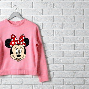 May include: A light pink sweatshirt featuring a Minnie Mouse graphic. The graphic shows Minnie's face, with black ears and a red bow with white polka dots. The sweatshirt is hanging on a gray hanger against a white brick wall.