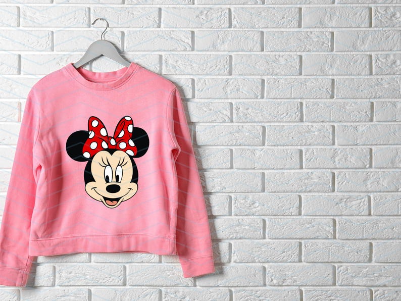 May include: A light pink sweatshirt featuring a Minnie Mouse graphic. The graphic shows Minnie's face, with black ears and a red bow with white polka dots. The sweatshirt is hanging on a gray hanger against a white brick wall.
