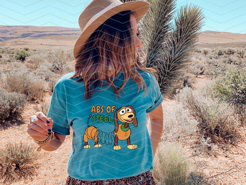 May include: Teal t-shirt featuring a cartoon image of Slinky Dog with the text "ABS OF STEEL". The design includes a brown dog with a spring body. The shirt is worn by a person in a desert setting.