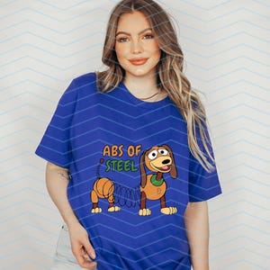 May include: A royal blue t-shirt featuring a cartoon image of Slinky Dog from Toy Story. The graphic includes the text "ABS OF STEEL" in orange and green. The design is centered on the shirt.