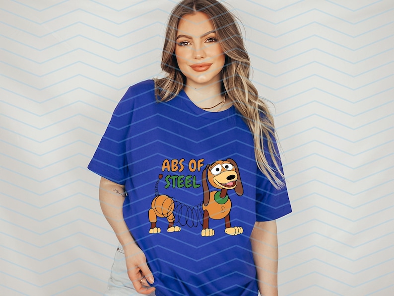 May include: A royal blue t-shirt featuring a cartoon image of Slinky Dog from Toy Story. The graphic includes the text "ABS OF STEEL" in orange and green. The design is centered on the shirt.