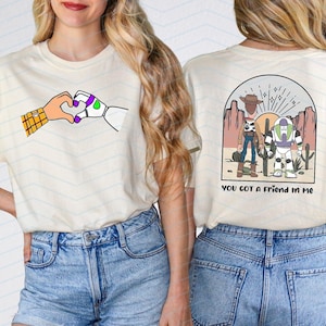 Pu&ograve; includere: T-shirt color crema con un'illustrazione a cartoni animati di due mani che formano un cuore. Una mano &egrave; un robot, l'altra un cowboy. Il retro della maglietta presenta una scena del deserto con personaggi dei cartoni animati e la scritta "YOU GOT A FRIEND IN ME."