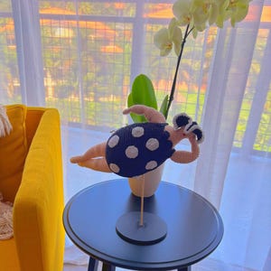 Può includere: Una figura all'uncinetto fatta a mano di una persona in costume da bagno, sdraiata. Il costume da bagno è blu navy con pois bianchi. La figura è esposta su un tavolo nero, con un'orchidea gialla sullo sfondo.