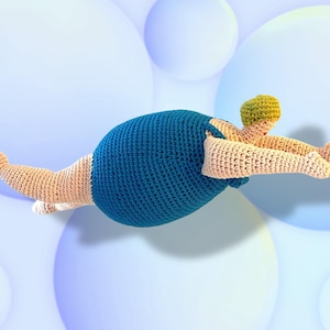 Può includere: Una figura all'uncinetto in posizione di nuoto, con un corpo blu, arti beige e una testa giallo-verde. La figura è posizionata su uno sfondo blu chiaro morbido con globi bianchi, creando un effetto stravagante.