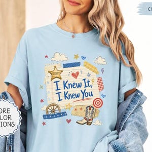 May include: A light blue t-shirt featuring a graphic print of a notepad page with the text "I Knew It, I Knew You". The design includes a sheriff star, a heart, a wagon wheel, a cowboy boot, and a target symbol in red, blue, and yellow colors.