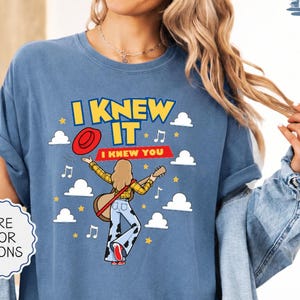 May include: A blue jean colored t-shirt featuring a graphic of a person with blonde hair playing a guitar, wearing a yellow shirt and cow print pants. The text reads I KNEW IT and I KNEW YOU, surrounded by white clouds, yellow stars, and musical notes.