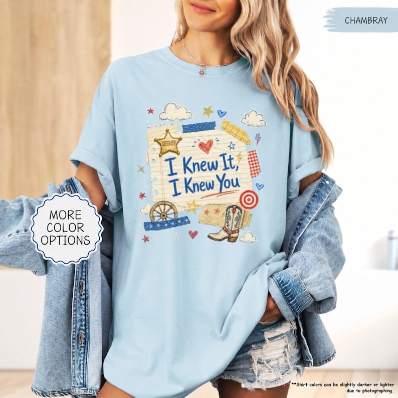 May include: A light blue t-shirt featuring a graphic print of a notepad page with the text "I Knew It, I Knew You". The design includes a sheriff star, a heart, a wagon wheel, a cowboy boot, and a target symbol in red, blue, and yellow colors.