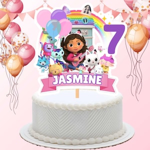 May include: A white frosted cake with a decorative topper featuring Gabby's Dollhouse characters, balloons, and the name "Jasmine". The number 7 is also displayed. Pink and gold balloons are in the background.